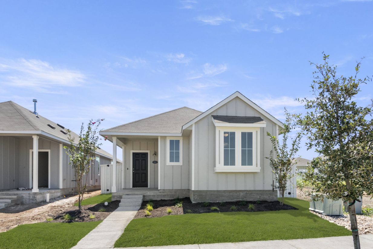 An image of gray single-story house with a dark blue door, grass landscape, trees, and blue sky in the background.