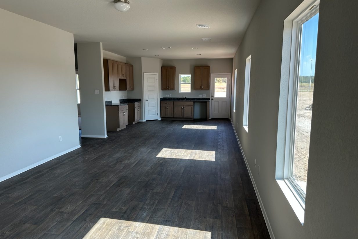 Image of home living areas with kitchen in rear. Wood-look flooring, white walls and windows down one wall, on rear door, and over sink.