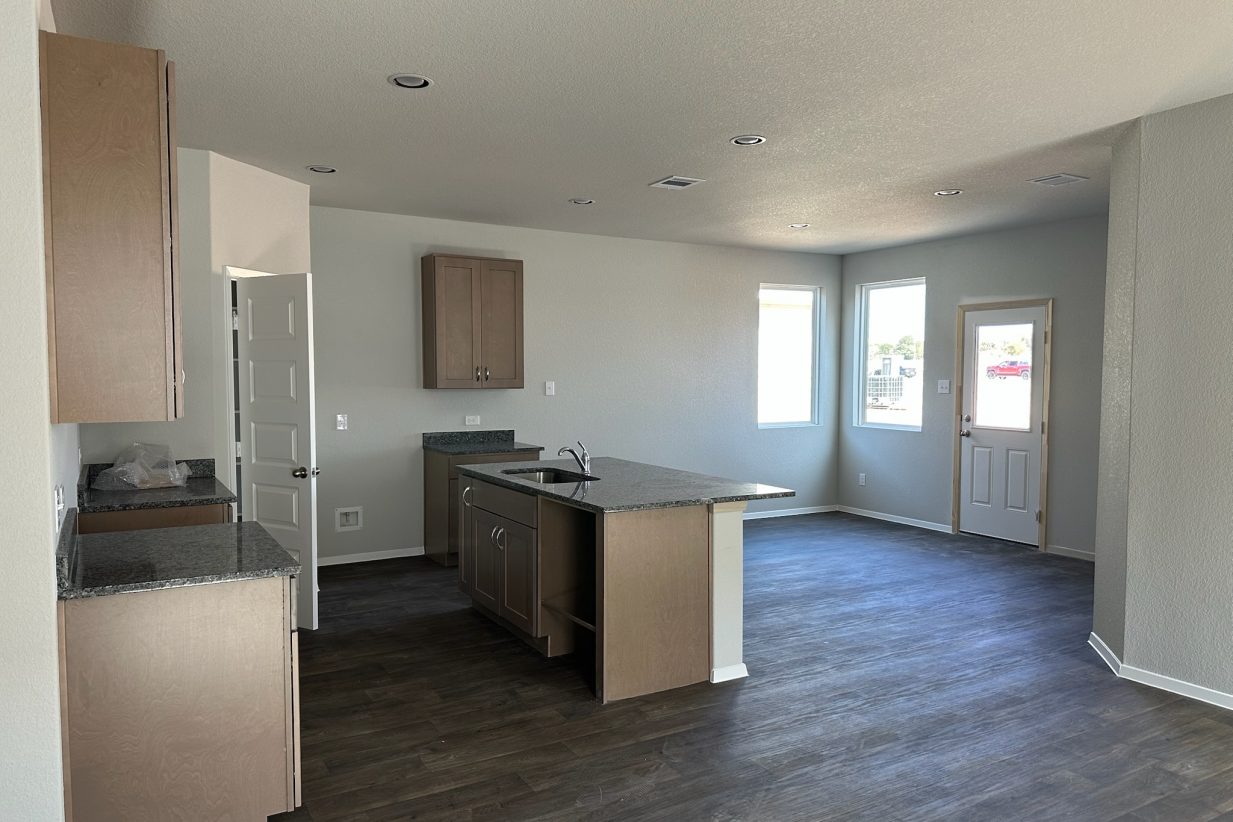 Image of home kitchen and dining room with light brown cabinets, center island, wood-look floors, and white walls with windows