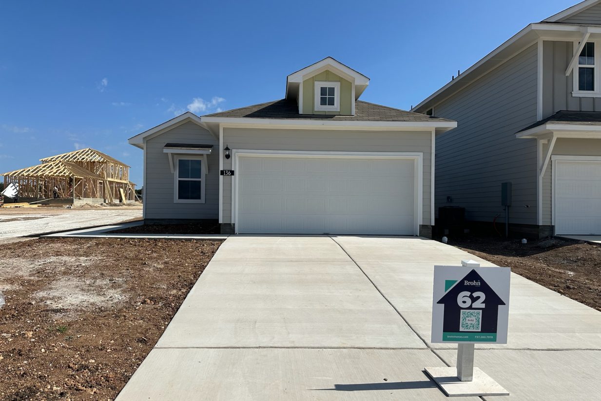 Image of one story home with tan siding, yellow accent, and white trim and garage door