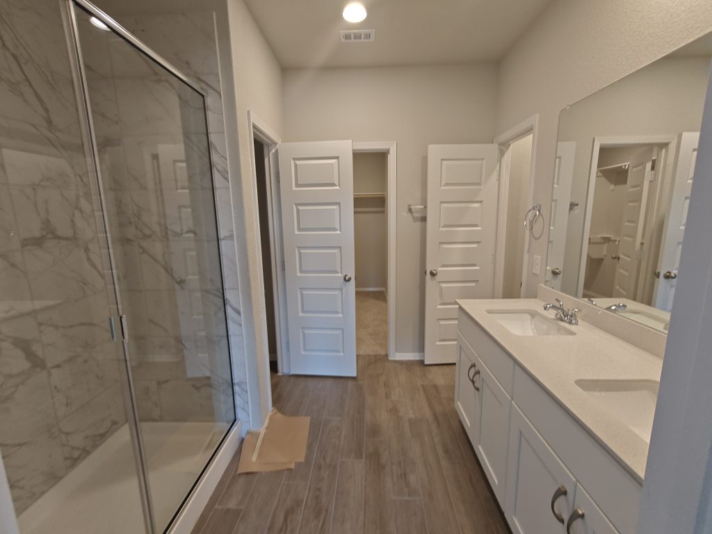 An image of a bathroom wood vinyl flooring, a walk-in glass shower with marble tiles, a dual vanity with silestone countertops with white cabinetry.