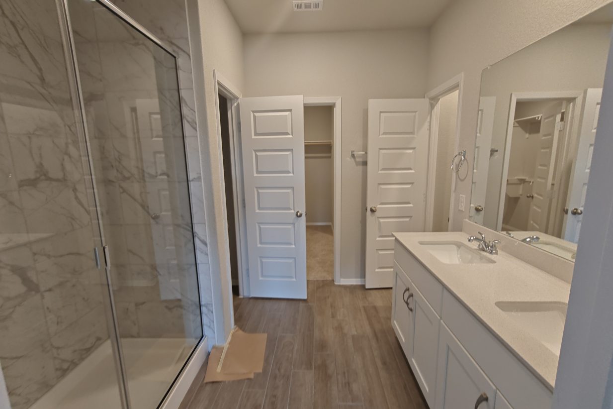 An image of a bathroom wood vinyl flooring, a walk-in glass shower with marble tiles, a dual vanity with silestone countertops with white cabinetry.