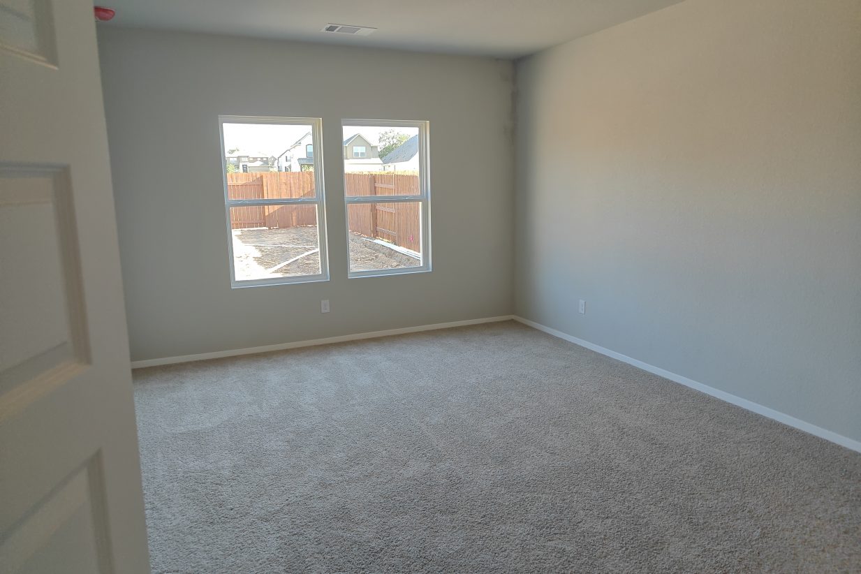 An image of the bedroom with white-framed windows, cream colored painted walls, and tan carpet flooring.