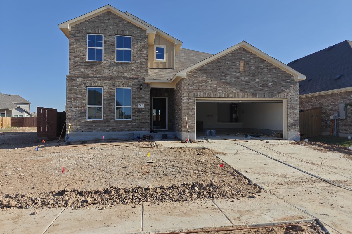 Image of the two-story brick home with an open garage, concrete driveway, and dirt landscape with a blue sky background.