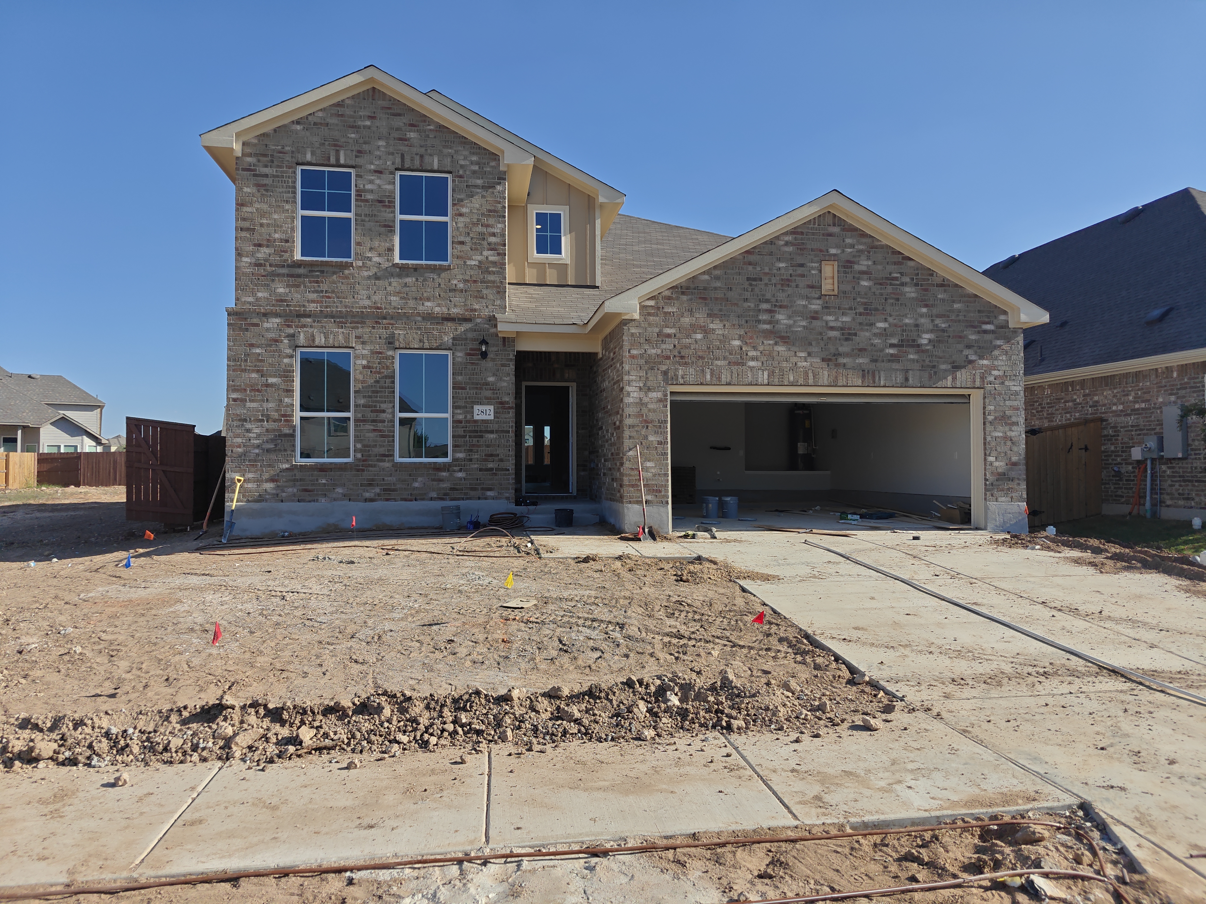 Image of the two-story brick home with an open garage, concrete driveway, and dirt landscape with a blue sky background.