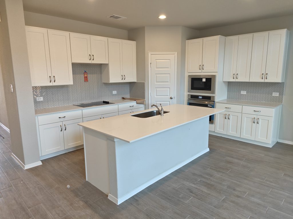 Image of a kitchen with white countertop and white cabinetry, grey blacksplash tile, an island, kitchen sink, steel