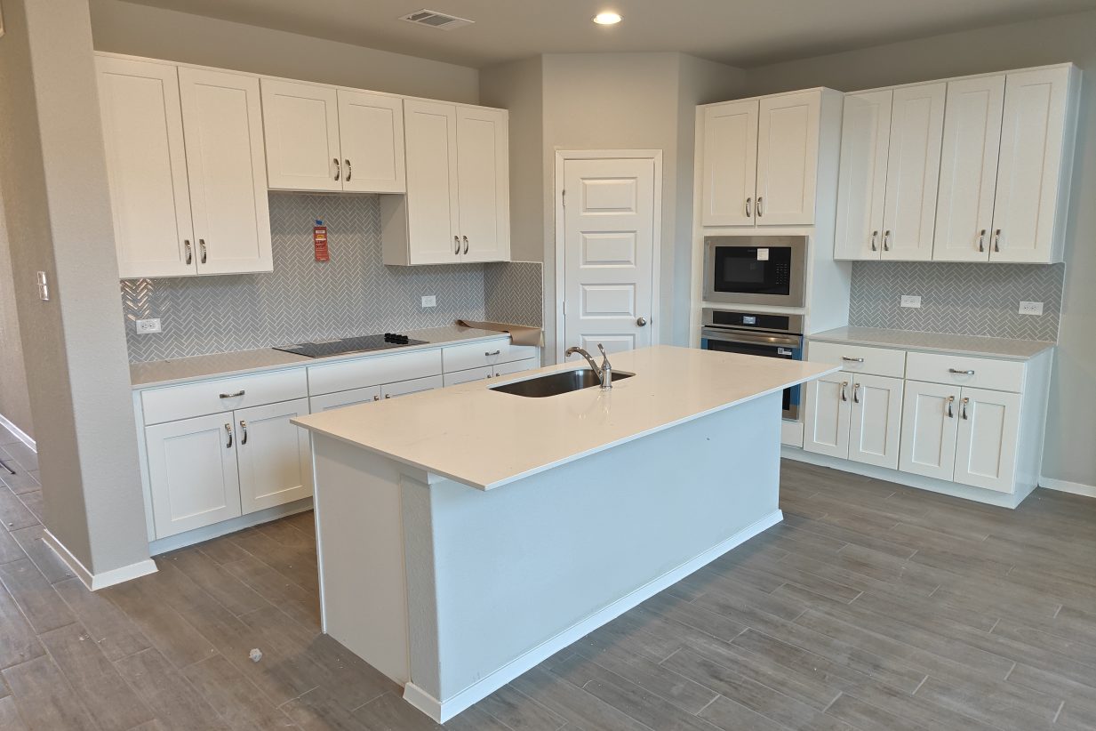 Image of a kitchen with white countertop and white cabinetry, grey blacksplash tile, an island, kitchen sink, steel