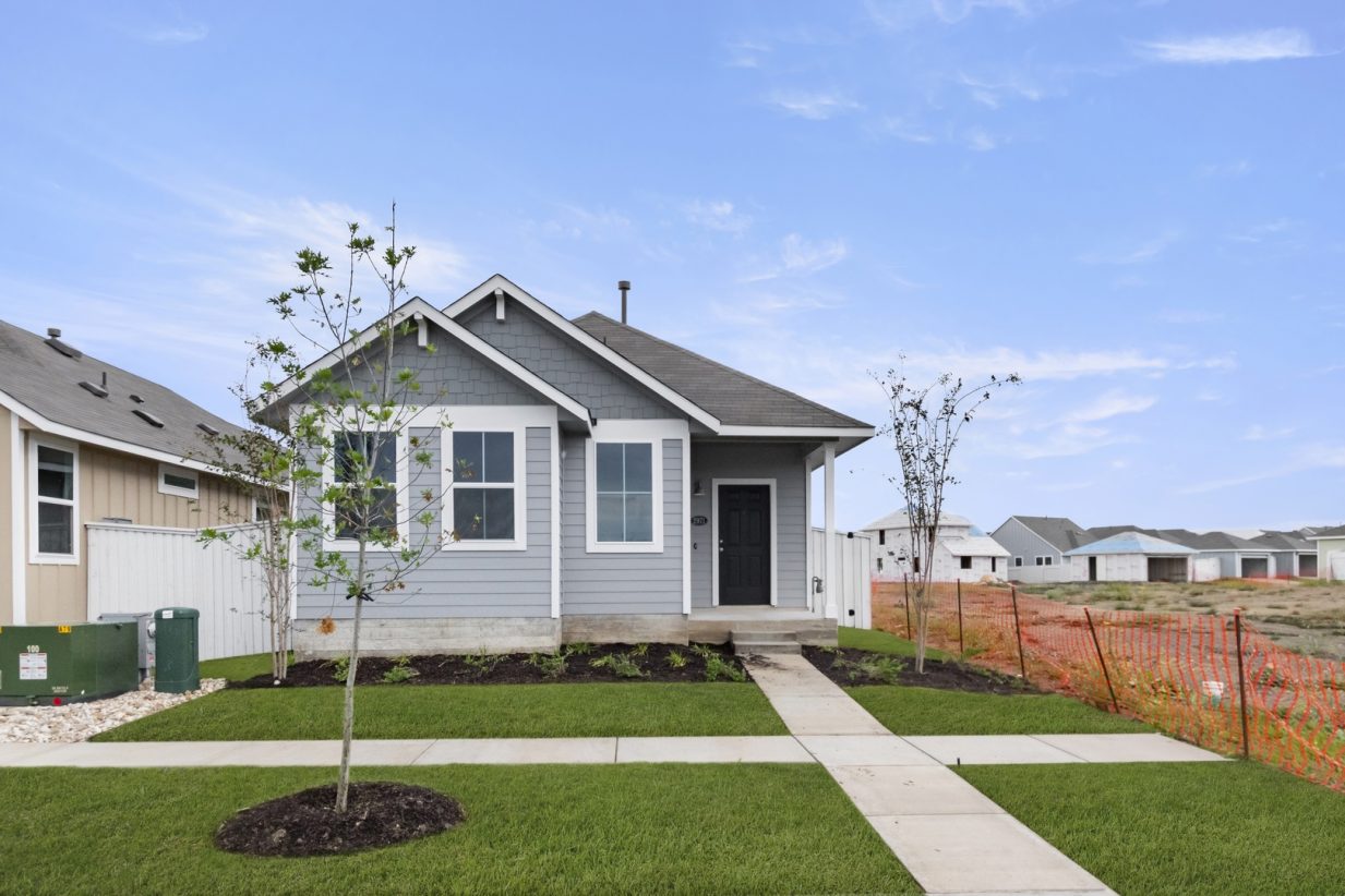 Image of exterior of a grey one story home with a black door and white trim with a tree and green grass and a blue sky