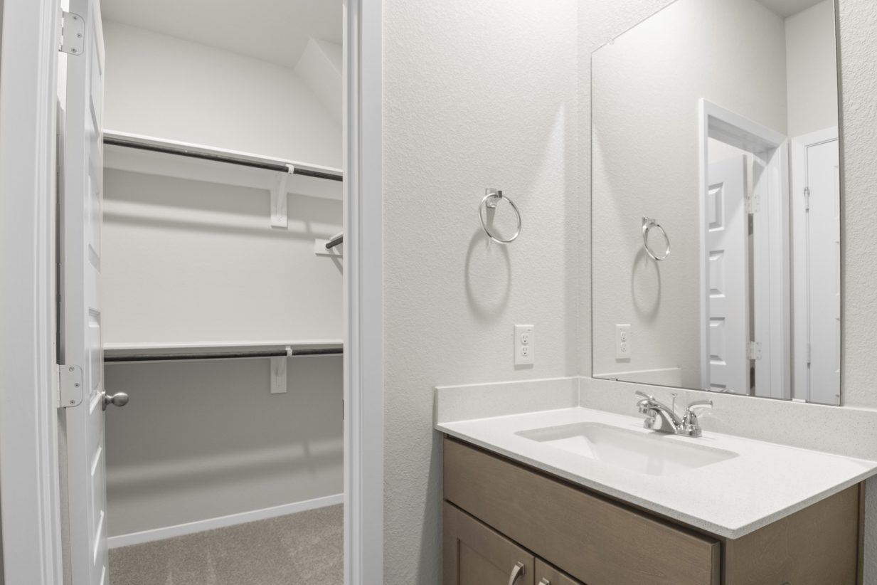 Image of a primary bathroom with brown cabinets and a white vanity with mirror and a connected walk-in closet