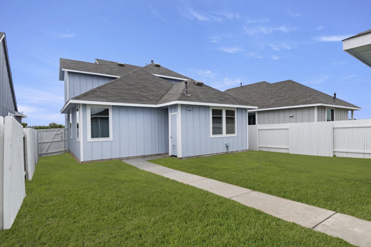 Image of the exterior of the back of a blue two story house with a cement pathway and a white fence with green grass and a blue sky