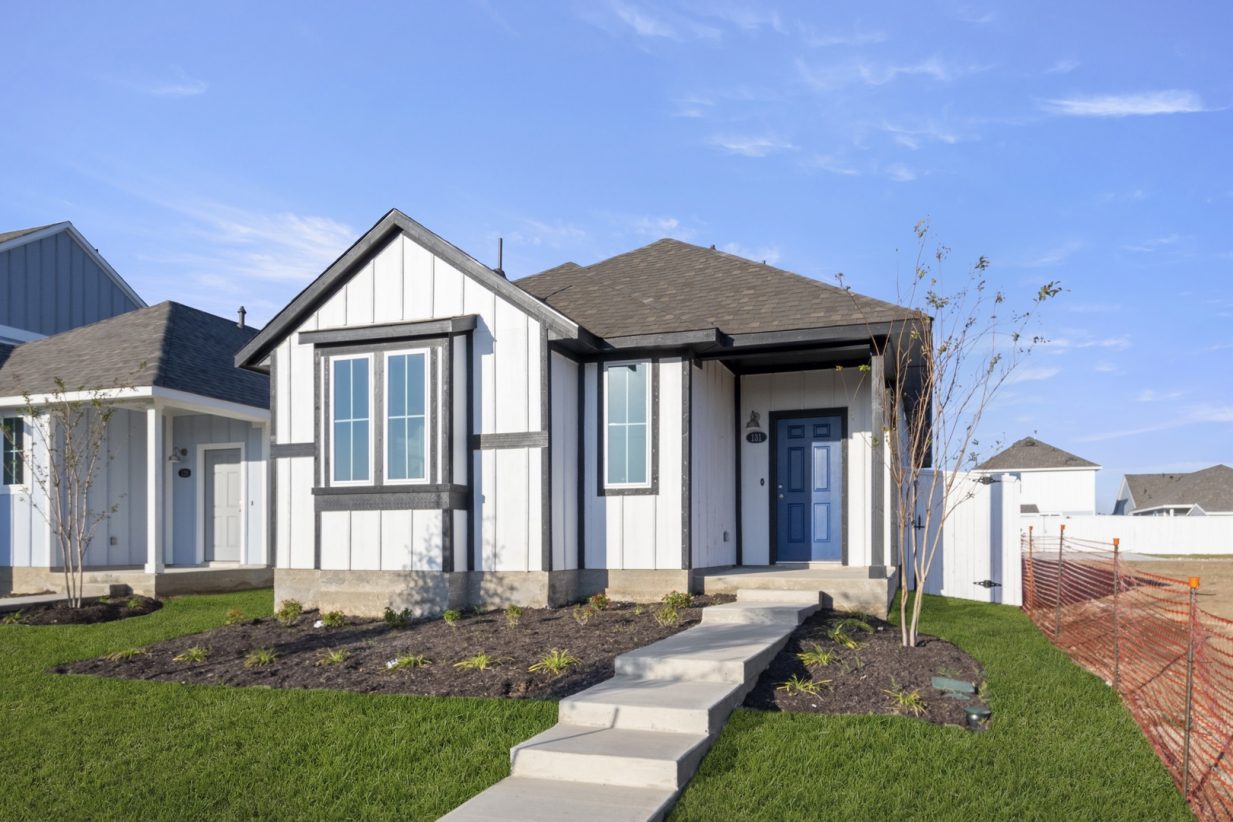Image of front of a white farmhouse-style single story home with black trimming with a green grass yard and blue sky in the background.