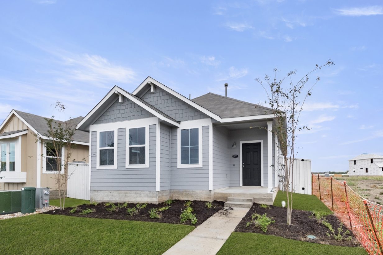 Image of exterior of a grey one story home with a black door and white trim with a tree and green grass and a blue sky