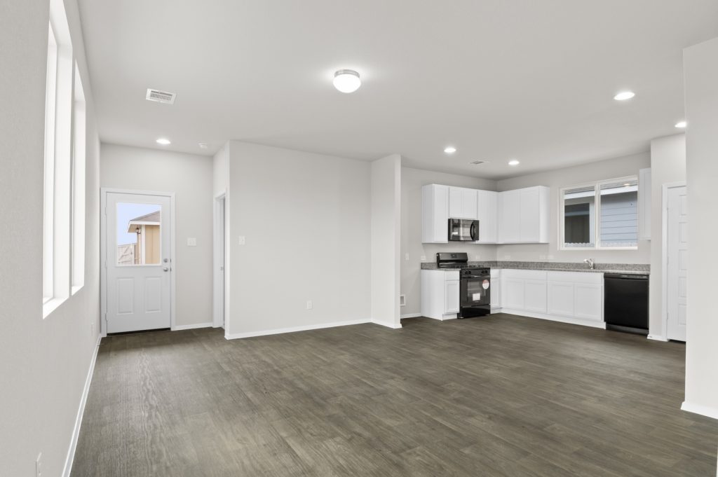 Image of a one story living room with brown flooring and grey walls and a kitchen in the distance