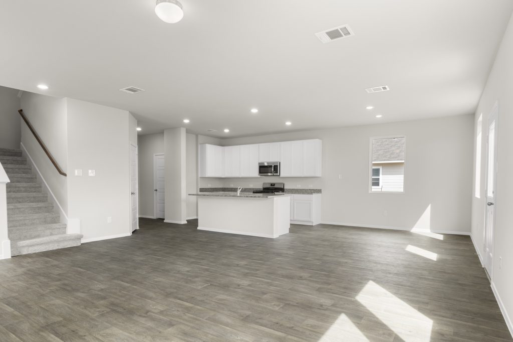 Image of two story home living room with brown flooring and light grey walls with a kitchen in the distance