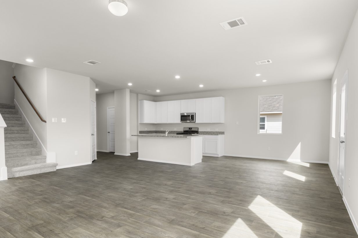 Image of two story home living room with brown flooring and light grey walls with a kitchen in the distance