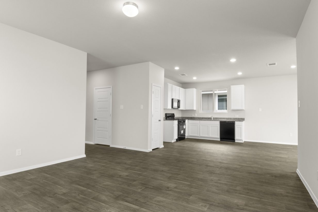 Image of a two story home living room with brown flooring and light grey walls and kitchen in the distance
