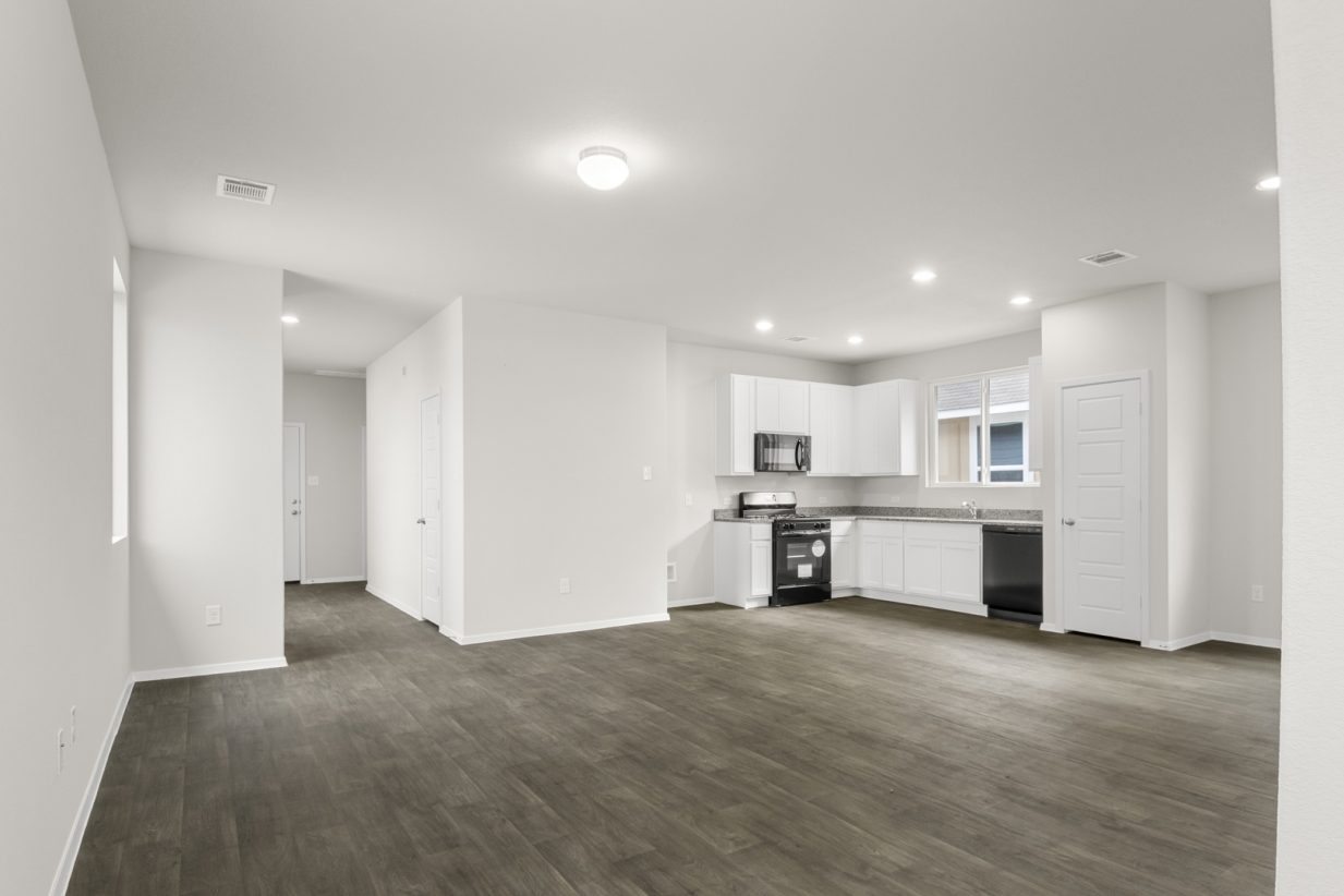 Image of living room with brown flooring and light grey walls with a kitchen in the distance