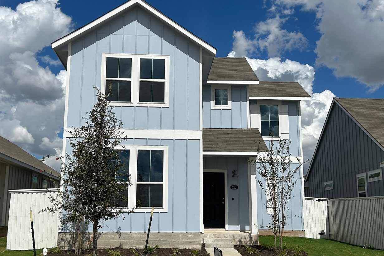Image of front exterior of a two-story blue house with a blue front door, green grass landscape, trees, and blue sky with clouds.