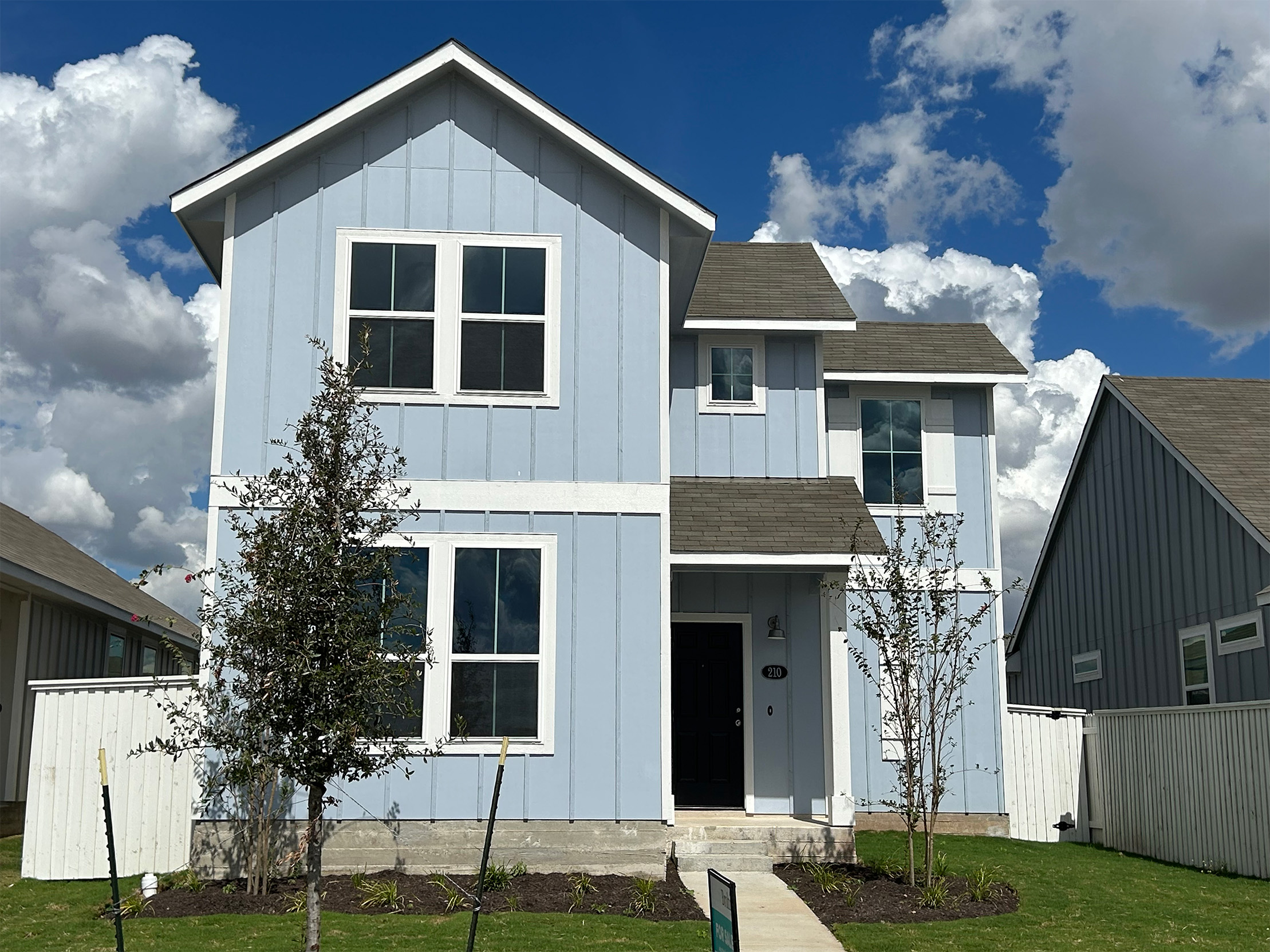 Image of front exterior of a two-story blue house with a blue front door, green grass landscape, trees, and blue sky with clouds.