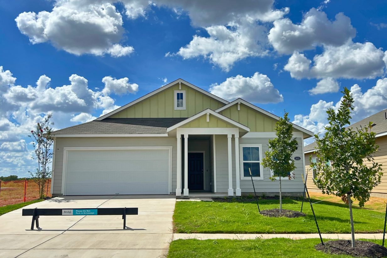 Image of front exterior of a single-story yellow house with white garage door, green grass landscape, and blue sky with clouds.