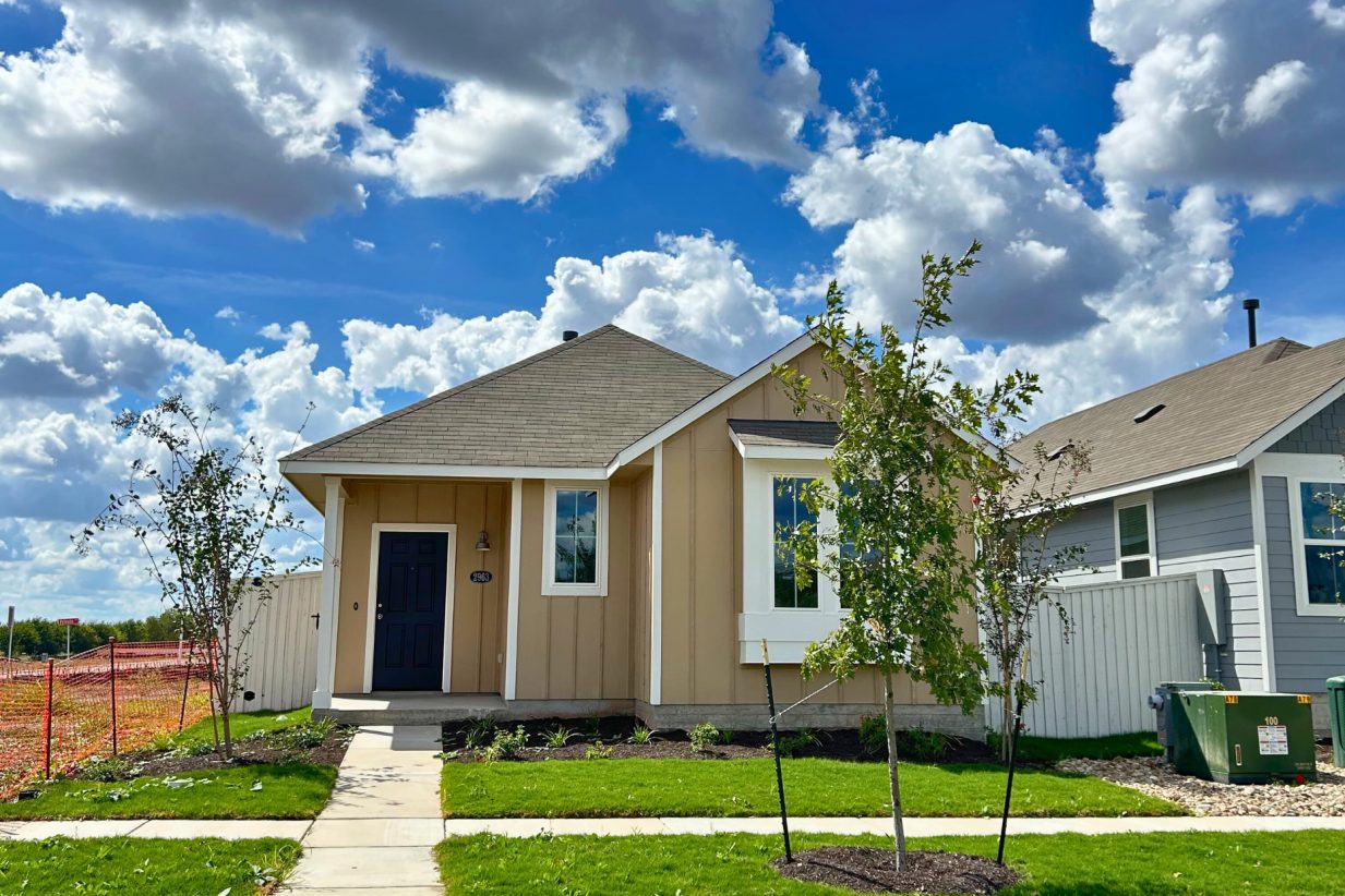 Image of front exterior of a single-story tan house with green grass landscape, trees, and blue sky with clouds.