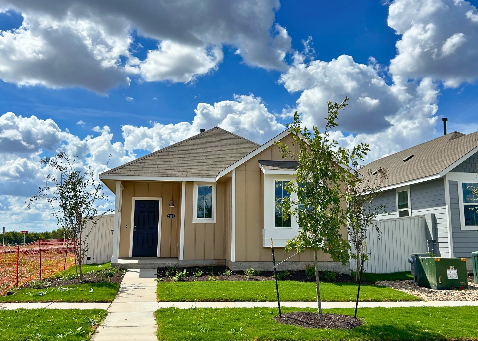 Image of front exterior of a single-story tan house with green grass landscape, trees, and blue sky with clouds.