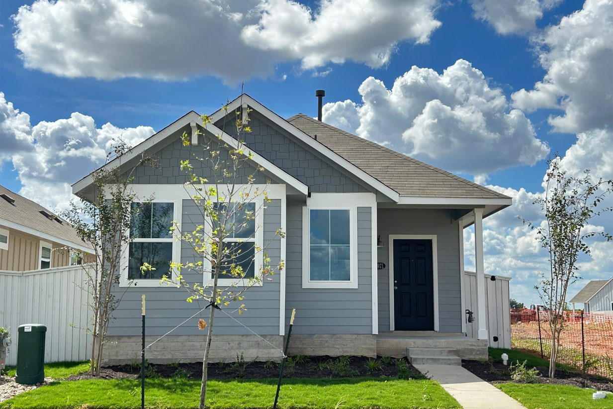 Image of front exterior of a single-story gray house with green grass landscape, trees, and blue sky with clouds.