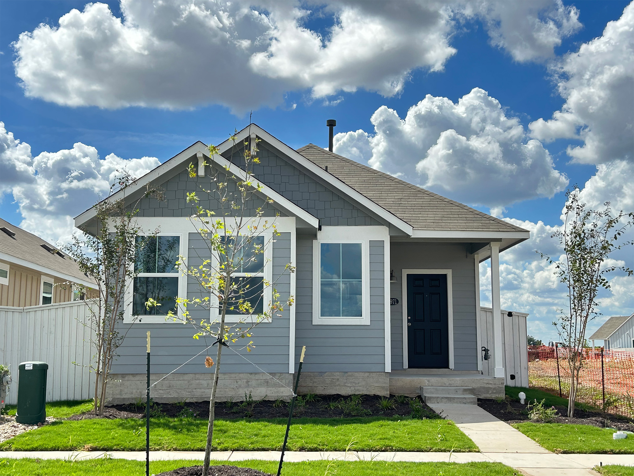 Image of front exterior of a single-story gray house with green grass landscape, trees, and blue sky with clouds.