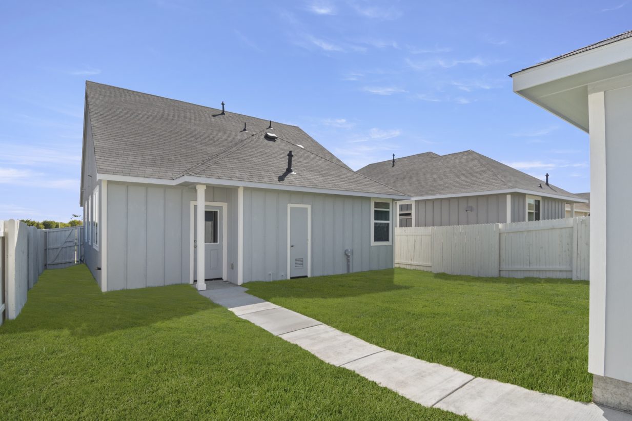 Image of back of blue one story house with walkway to detached garage, green grass, white fence, and sky
