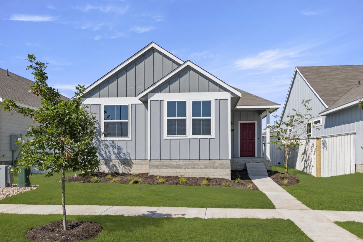 Image of front exterior of one-story grey home with red door, sidewalk, green grass, and sky