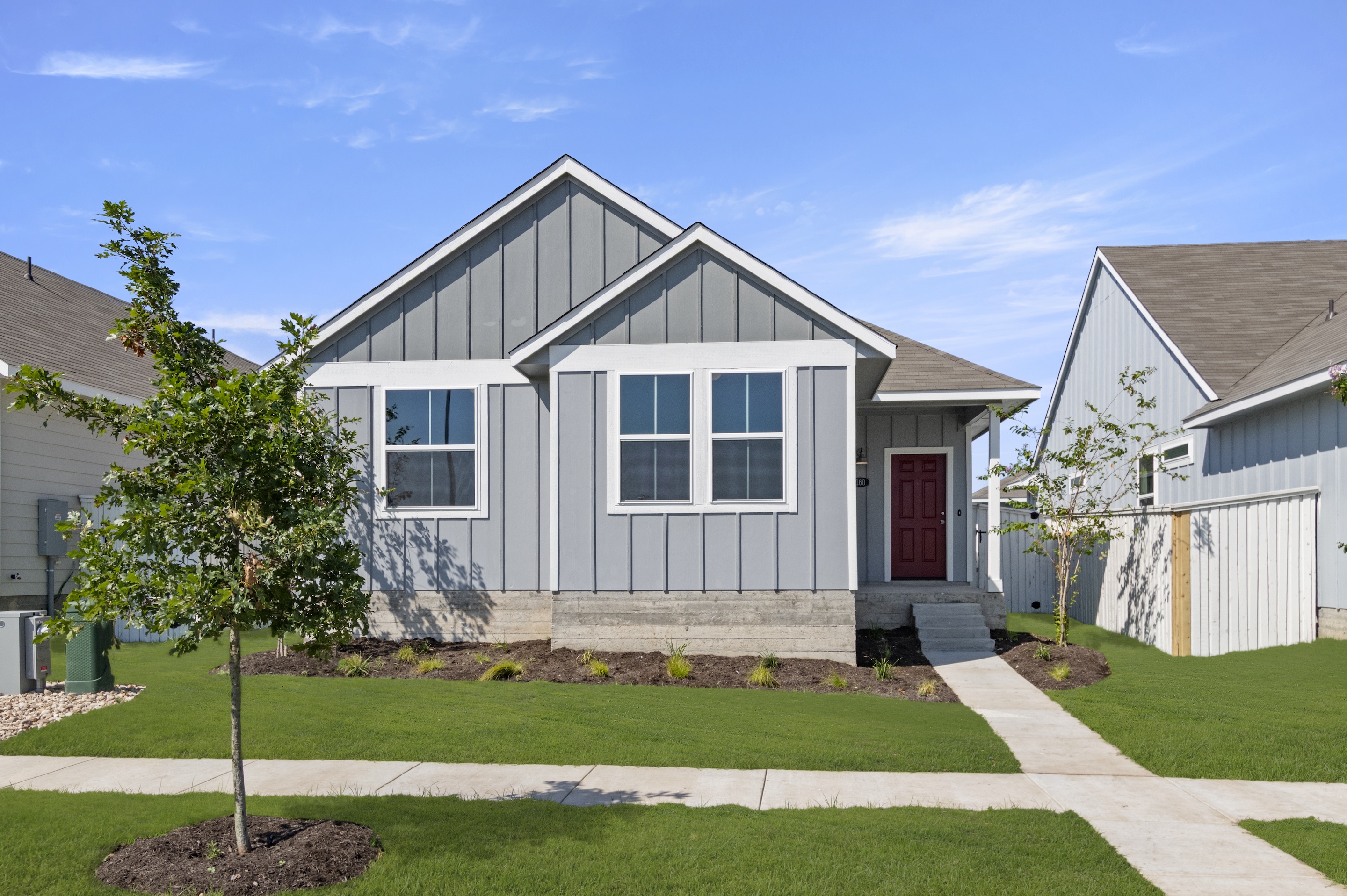 Image of front exterior of one-story grey home with red door, sidewalk, green grass, and sky