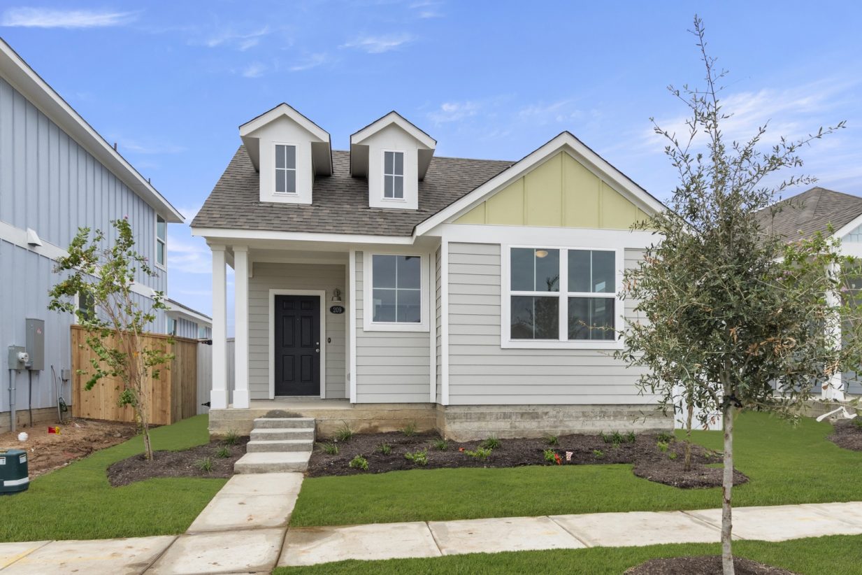 Image of one story home with grey siding and yellow accent siding, white trim, and black door. Path to front door, landscaping, and blue sky