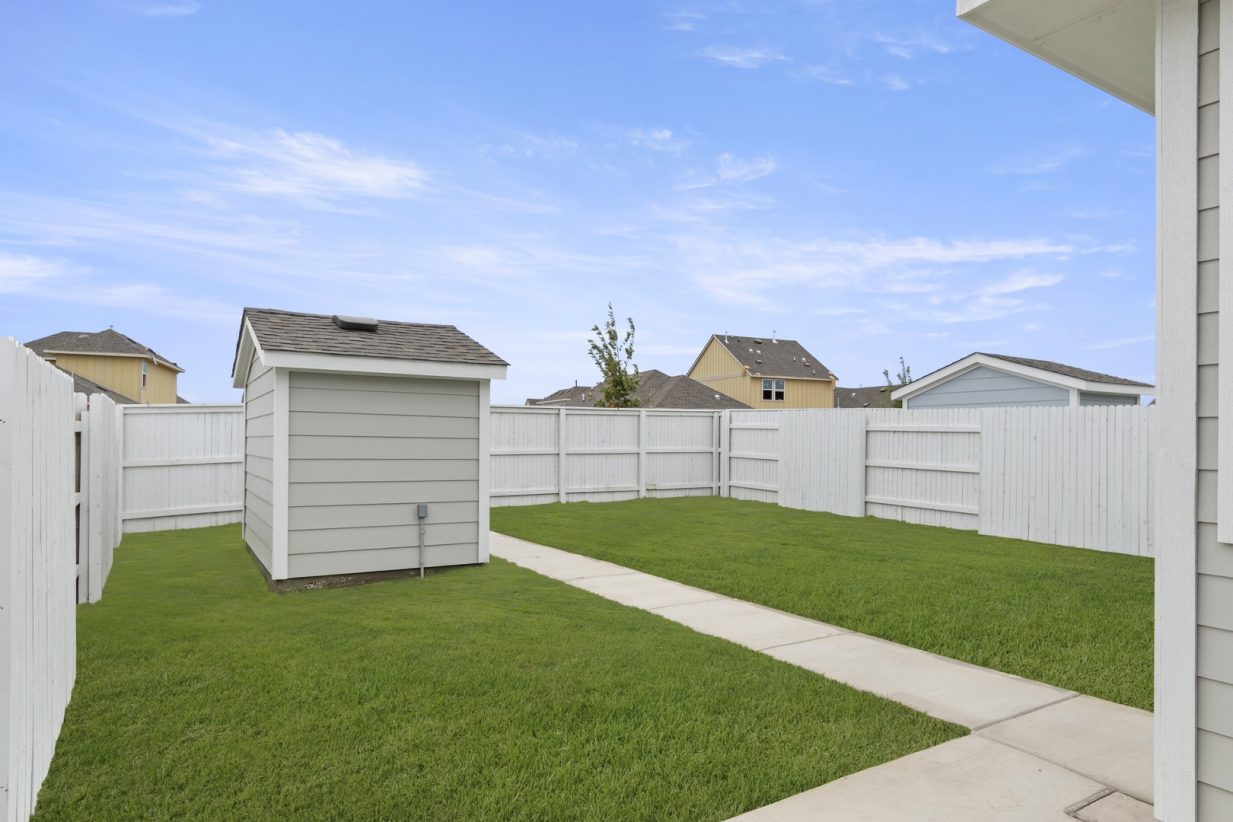 Image of home backyard with path to shed, white fence, green grass, and blue sky