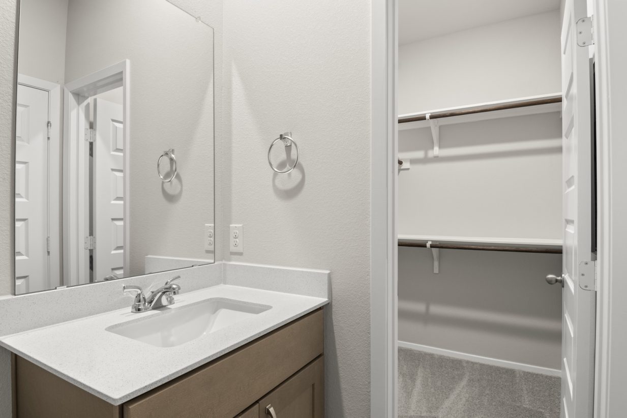 Image of home primary bathroom with sink with white countertop and light brown cabinet, mirror, and doorway to walk-in closet