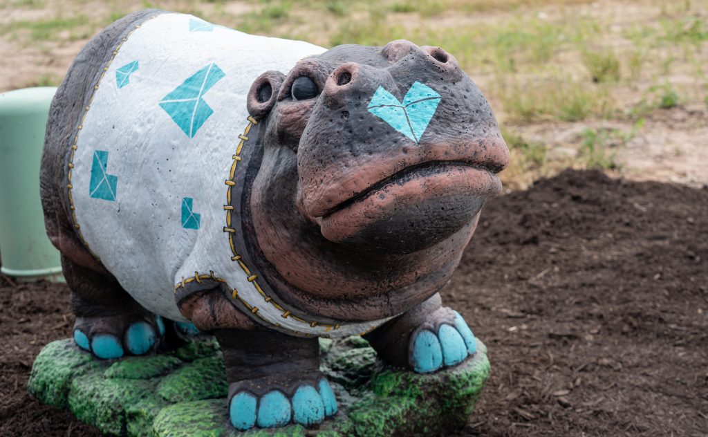 Image of a realistically painted hippo statue with a white t-shirt with teal hearts, teal nails, and a teal heart on the nose standing on painted grass