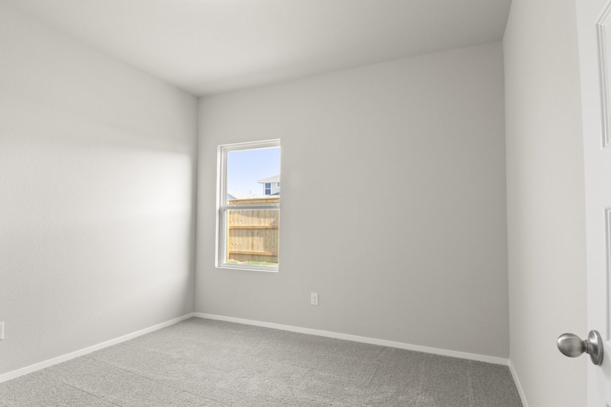 Image of a bedroom with tan carpeting and light grey walls with a window