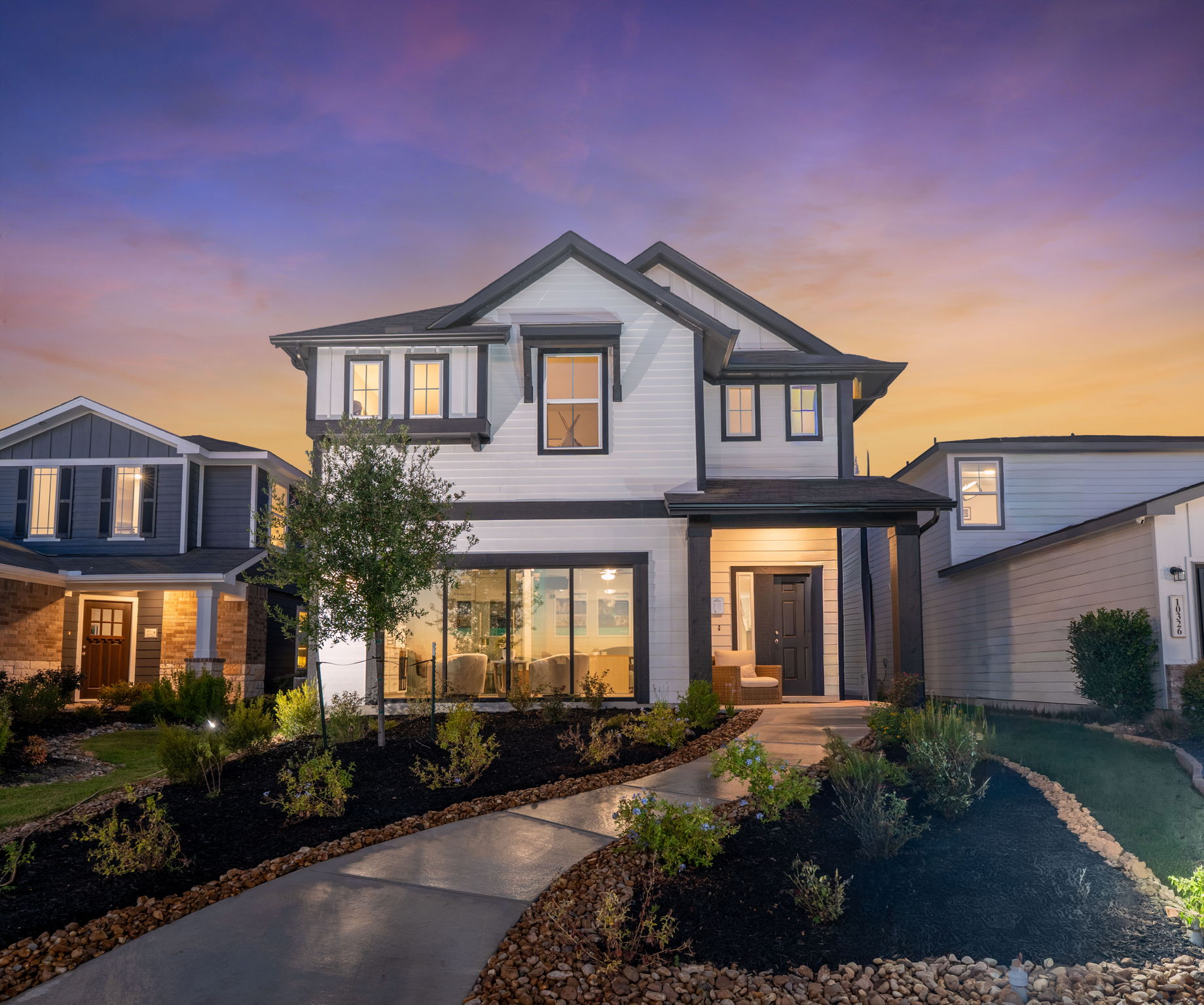 An exterior image a two-story white house with black framing, windows with light coming out front the outside, a concrete walk way, greenery with dirts, with a purple, blue, and yellow sunset in the background.