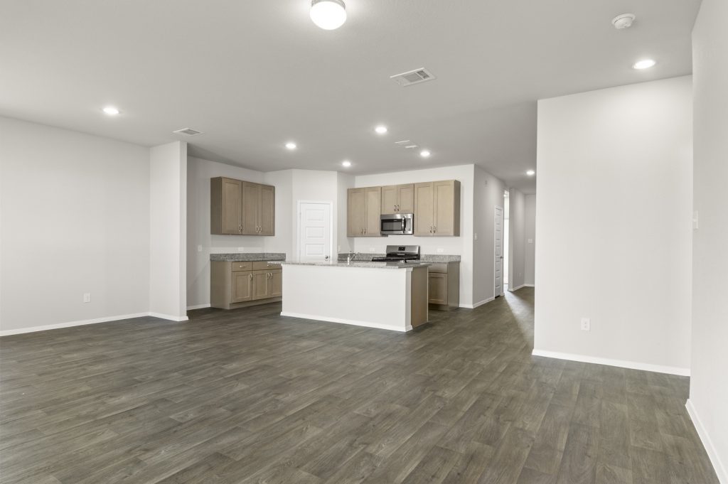 Image of a living room with dark vinyl flooring, cream walls and a kitchen in the distance