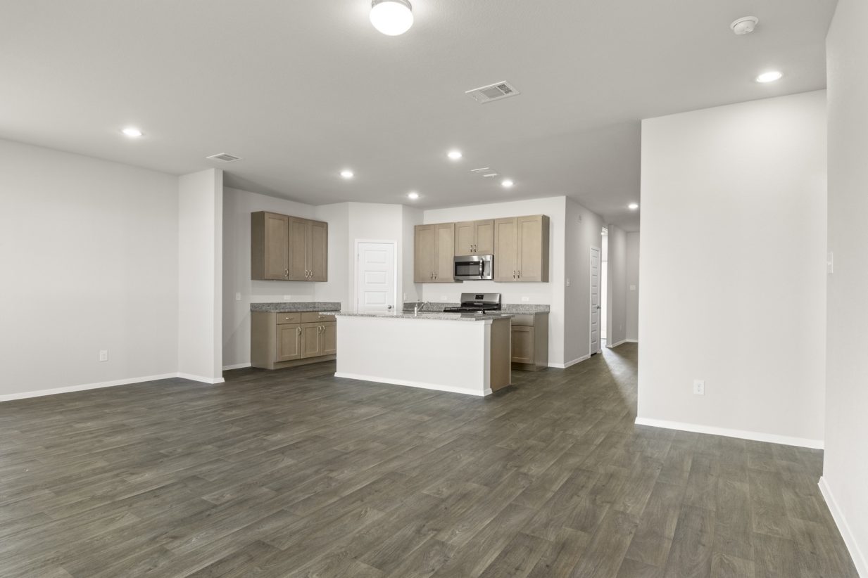 Image of a living room with dark vinyl flooring, cream walls and a kitchen in the distance