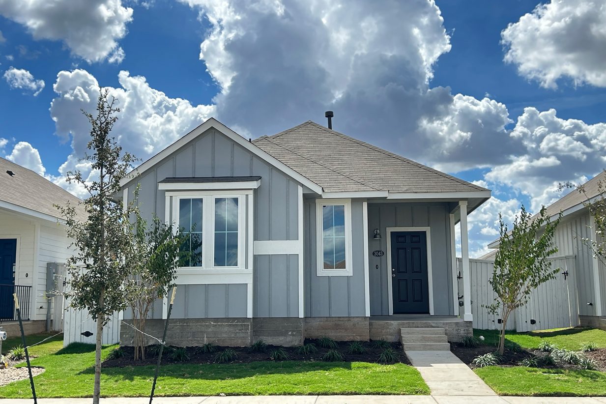Image of front exterior of a single-story gray house with a blue front door, green grass landscape, trees, and blue sky with clouds.