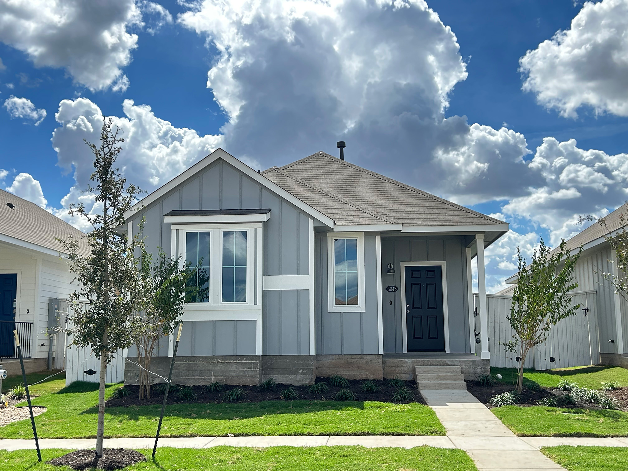 Image of front exterior of a single-story gray house with a blue front door, green grass landscape, trees, and blue sky with clouds.