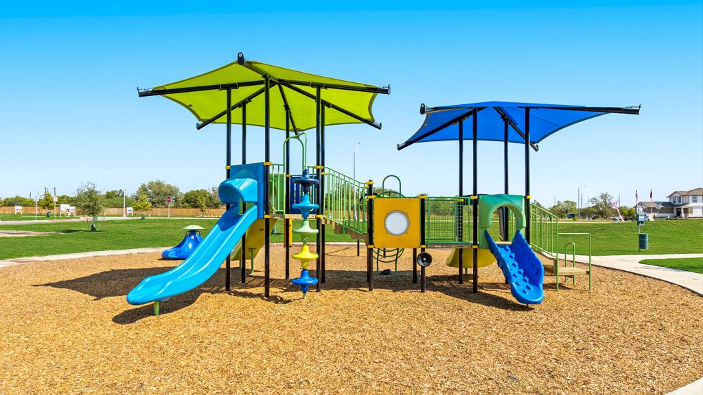 Image of community playground with green and blue fixtures and kid mulch. Open space behind and homes