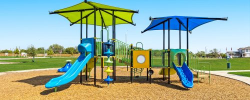 Image of community playground with green and blue fixtures and kid mulch. Open space behind and homes