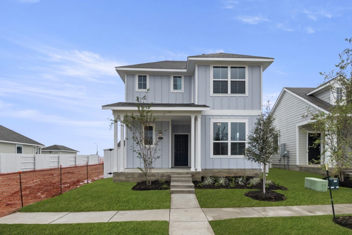 Image of exterior two story grey home with a black door, trees, green grass and a blue sky