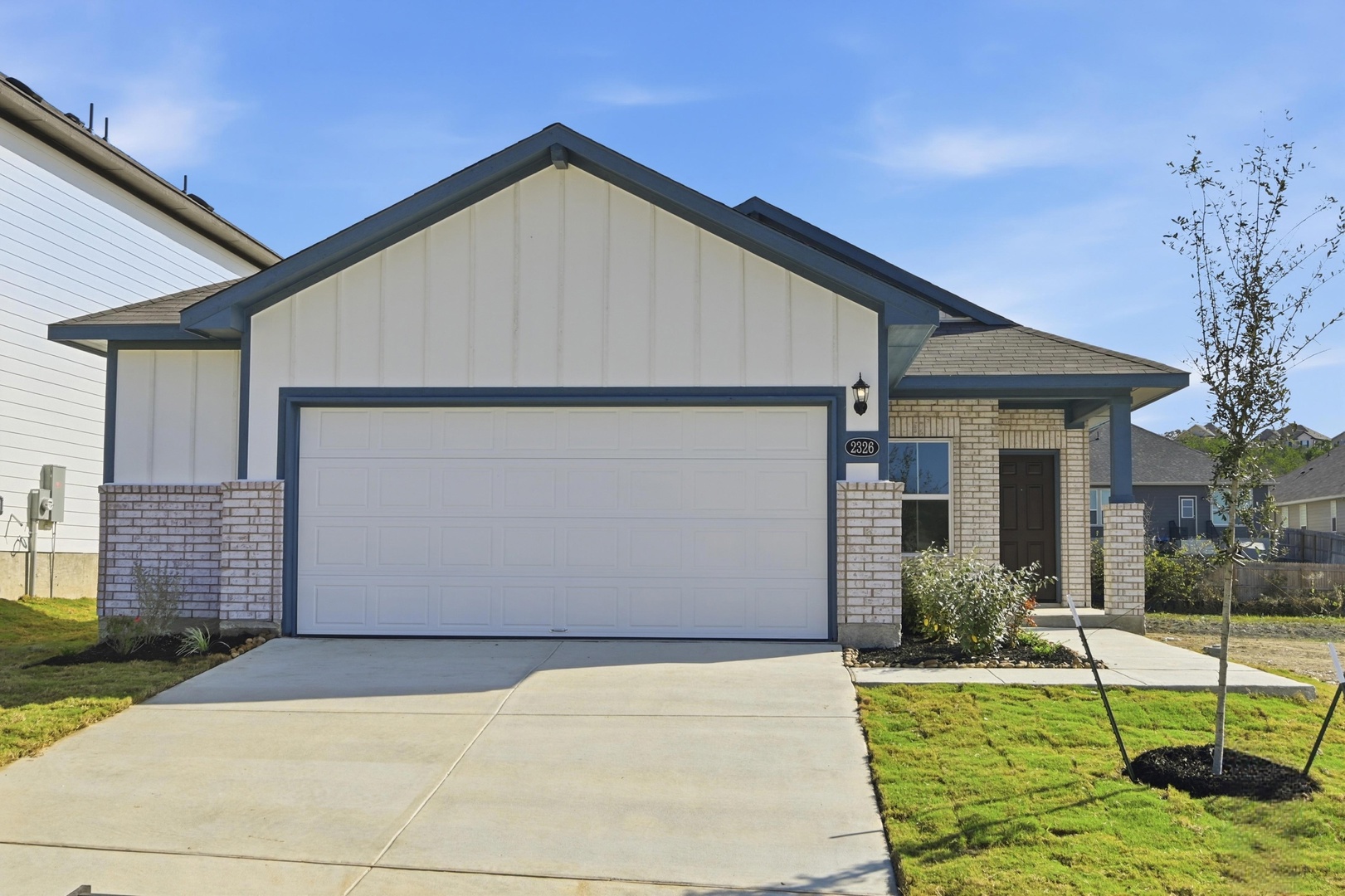 Image of the front exterior of a one story home with a white garage, brick accents, blue trim, a brown front door and a cement driveway