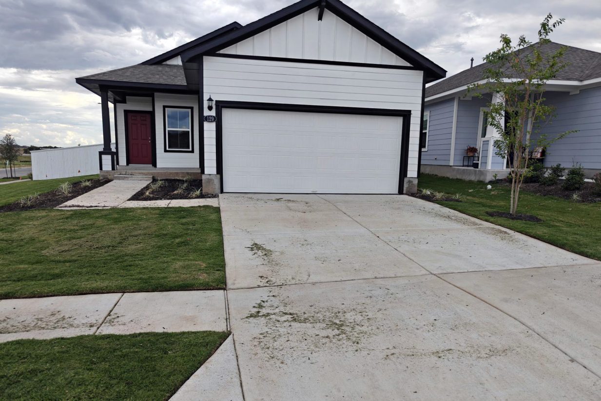 Image of one-story farmhouse style white home with black trim and red front door with driveway, green grass, and sky