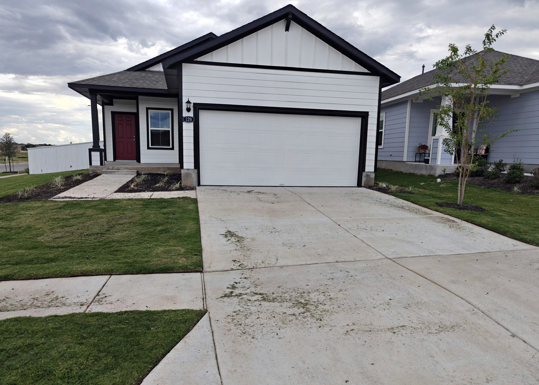 Image of one-story farmhouse style white home with black trim and red front door with driveway, green grass, and sky