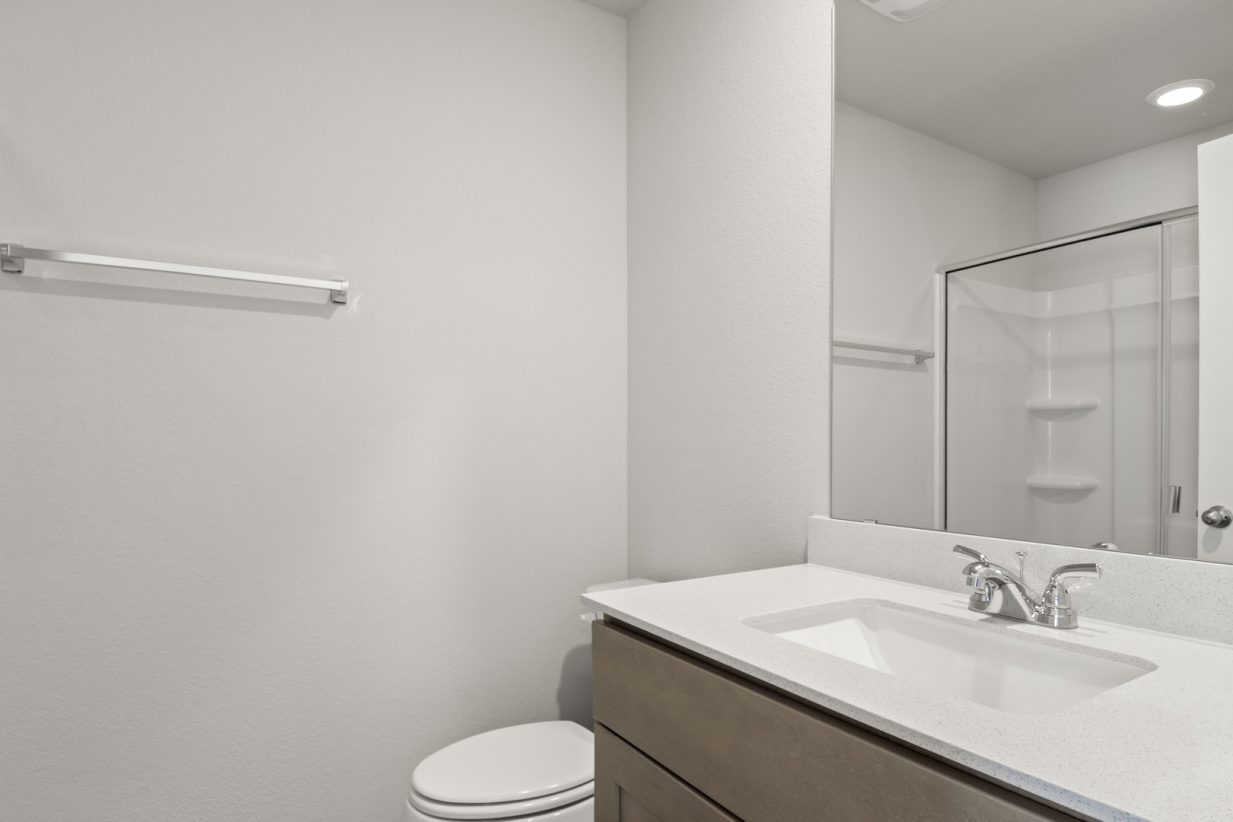 Image of a primary bathroom with light grey walls with brown cabinets, a white vanity and a mirror