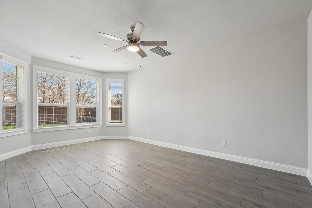Image of a primary bedroom with bay windows, light grey walls, dark wood flooring, a ceiling fan and white trim