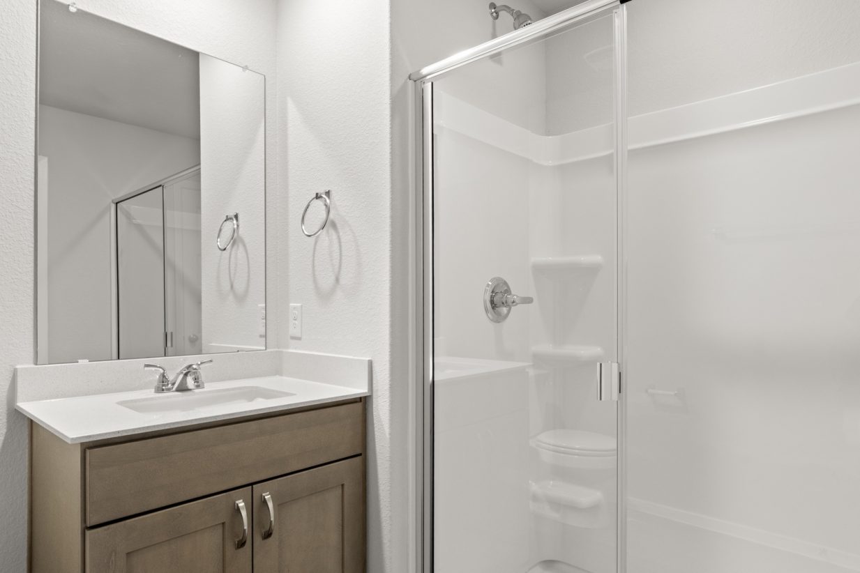 Image of a primary bathroom with a white vinyl walk-in shower, brown cabinets, and a white vanity with a mirror